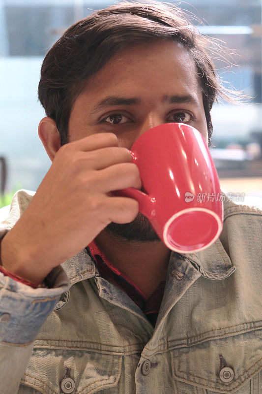 英俊的印度男子留着胡子，在酒店餐厅微笑着，一边吃着早餐，一边喝着红杯咖啡，卡布奇诺/茶/热巧克力，浅蓝色牛仔夹克